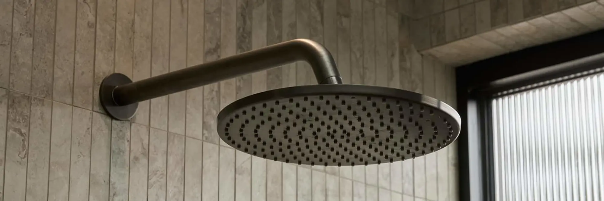 Modern round rainfall shower head with gunmetal finish mounted on tiled bathroom wall next to a window.