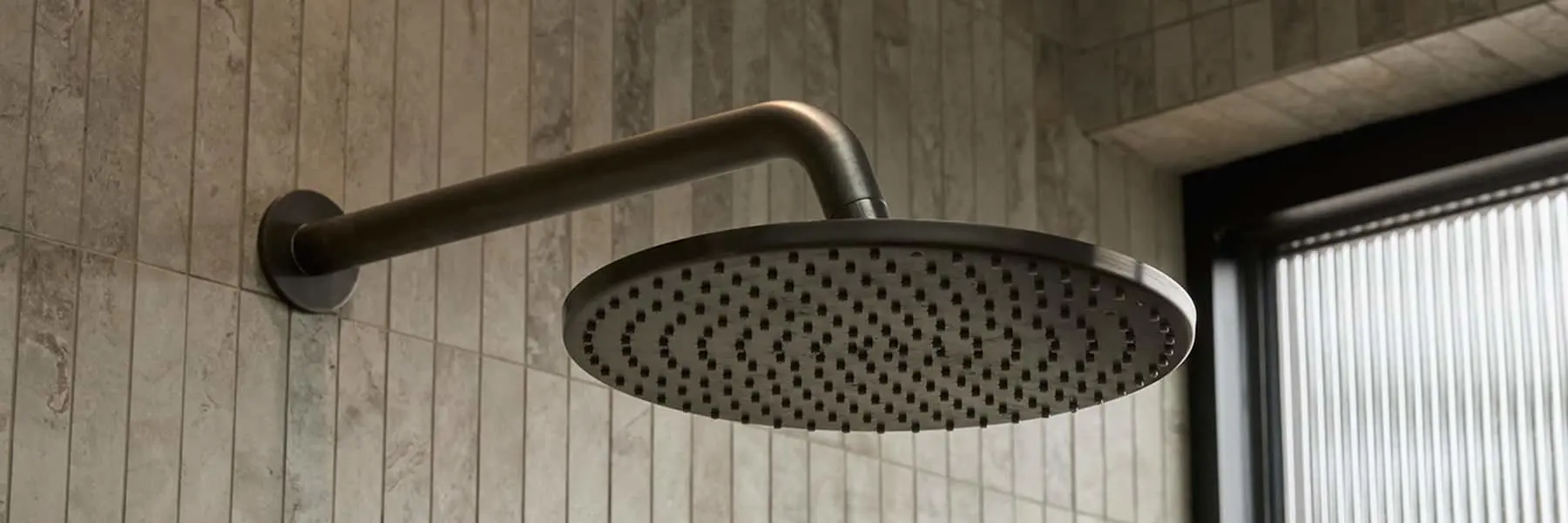 Modern round rainfall shower head with gunmetal finish mounted on tiled bathroom wall next to a window.