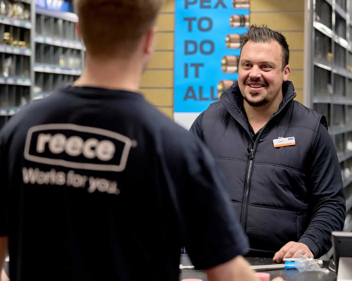 Two store employees in conversation at a hardware store counter, with shelves of products and a blue sign in background.