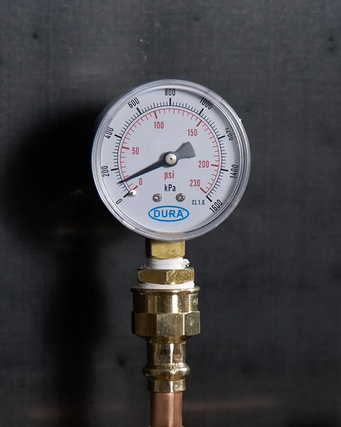 Pressure gauge with dual PSI/kPa readings mounted on brass fitting with copper pipe against dark background.