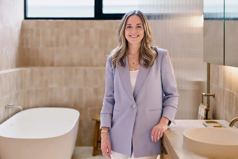 Person in light purple blazer smiling in a modern bathroom with beige tiles, white bathtub, and minimalist sink.