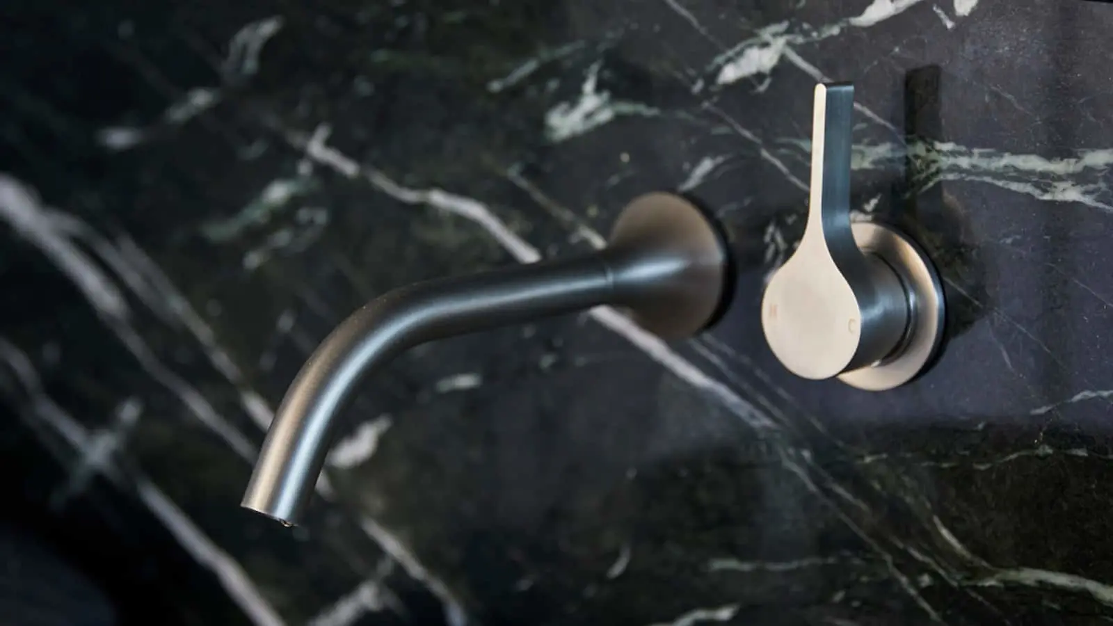 Modern wall-mounted bathroom faucet with curved stainless steel spout and minimalist handle against black marble.