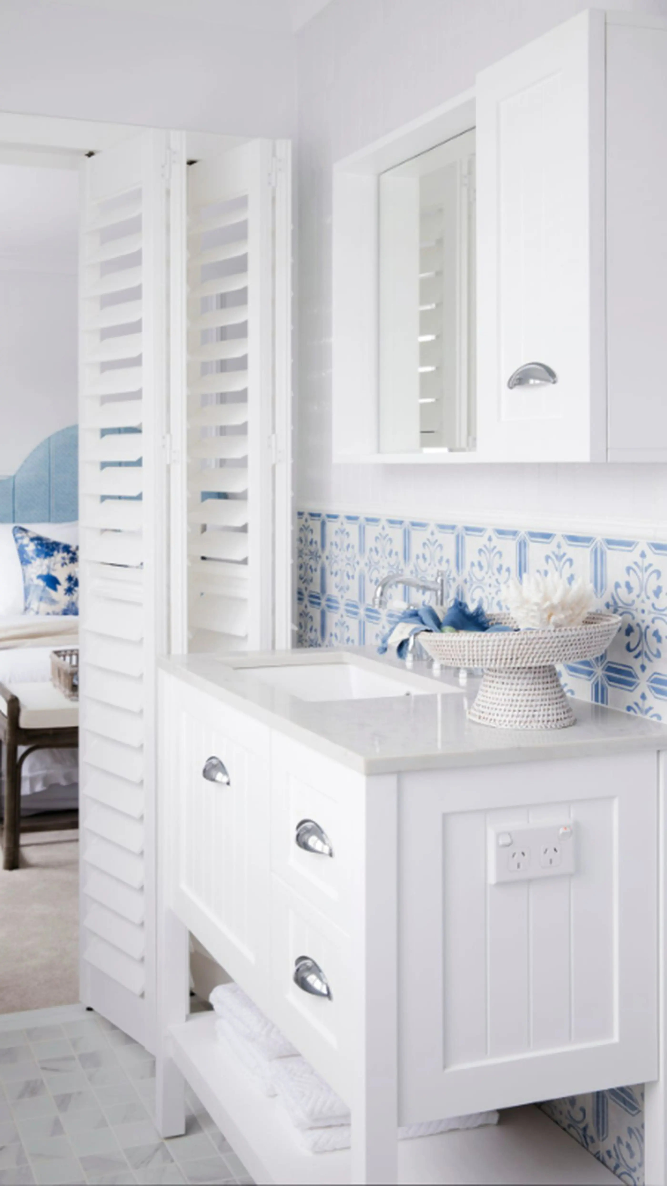 White bathroom vanity with blue and white tiled backsplash, louvered divider, and woven basket containing decorative items.