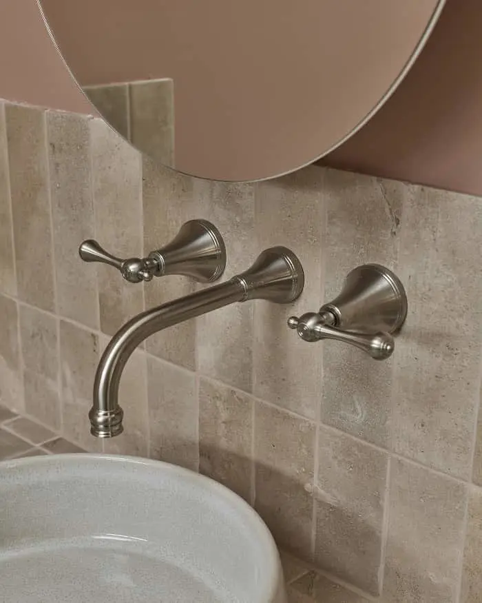 Wall-mounted brushed nickel bathroom faucet with two handles above a white sink, against beige tile backsplash.