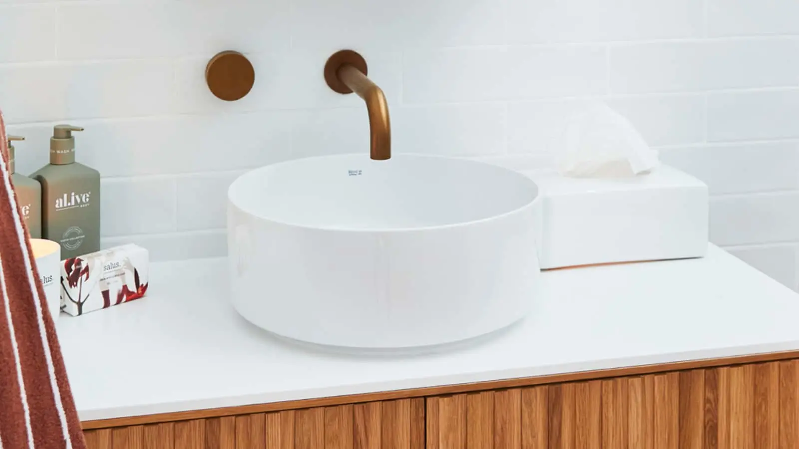 Modern bathroom with white circular vessel sink, bronze faucet, wooden vanity, and toiletries on white countertop.