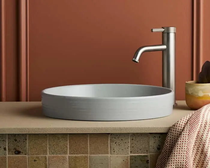 Modern bathroom sink with gray circular basin, brushed metal faucet, and small plant against terracotta wall.