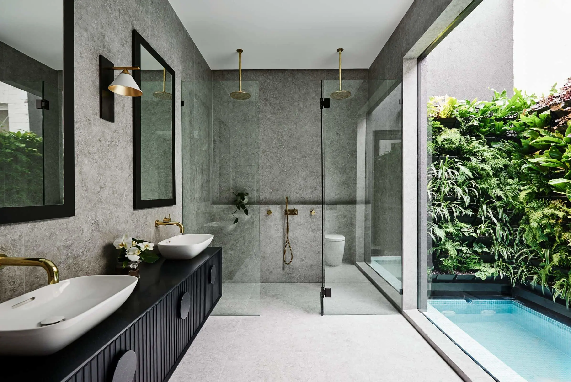 Modern bathroom with gray stone walls, dual sinks, glass shower, and large window overlooking a vertical garden and pool.