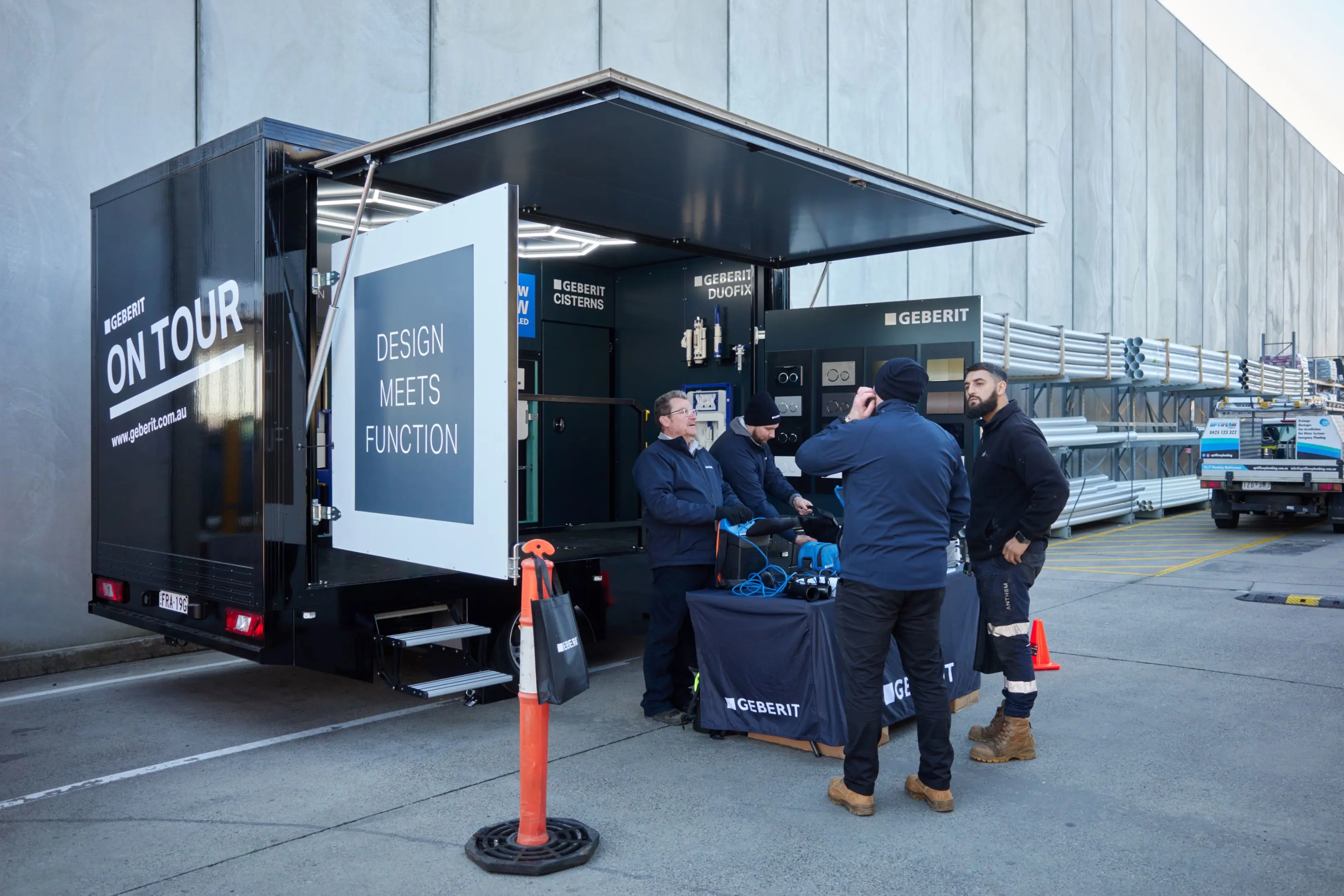 Four people gathered around a Geberit branded mobile showroom trailer parked outdoors, displaying plumbing products.