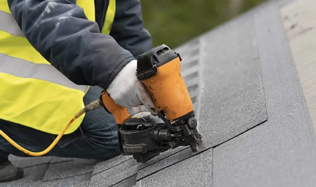 Construction worker in safety vest using orange nail gun to secure shingles on gray roof.