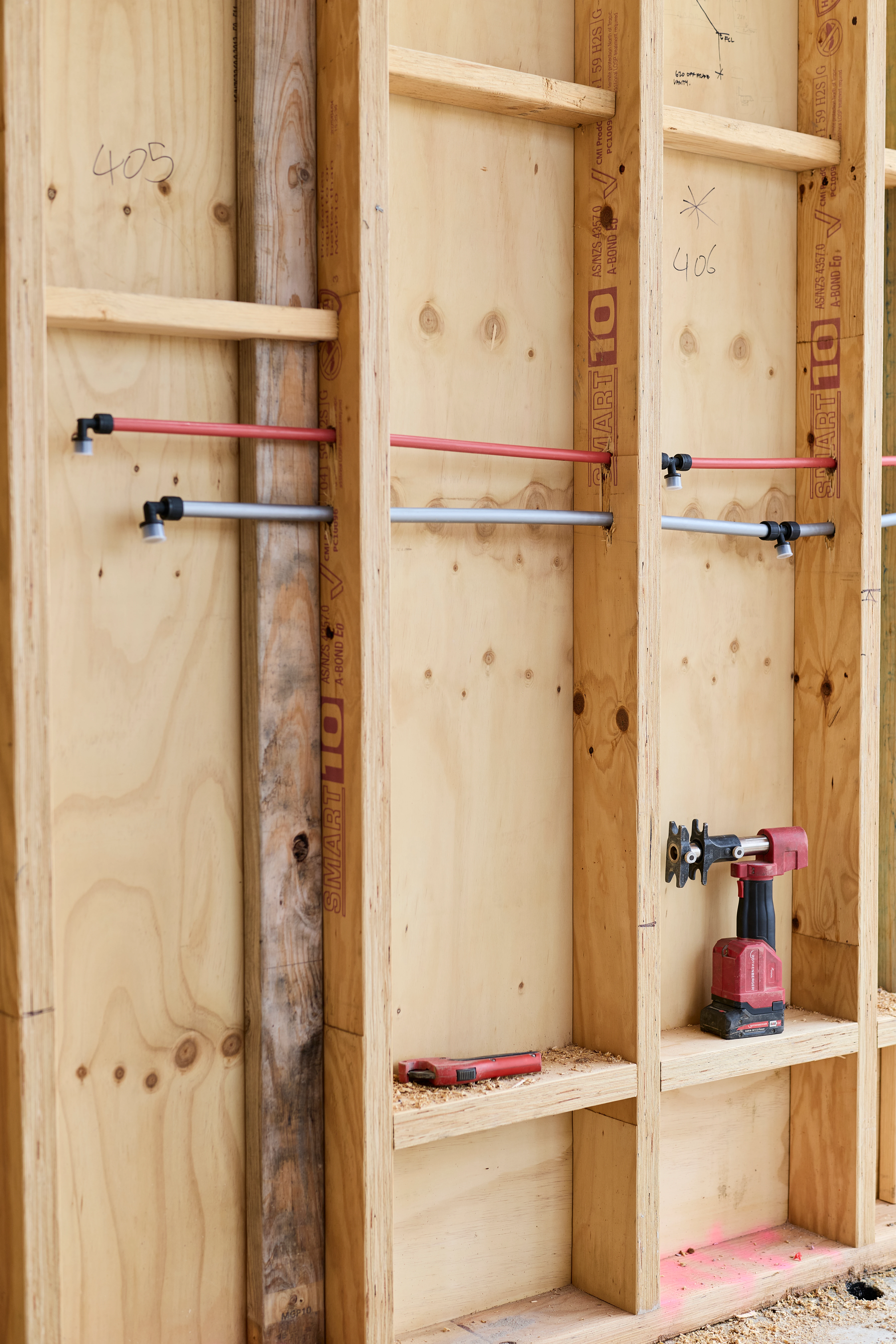 Wooden wall framing with plumbing pipes installed and a power drill resting in one section, marked with room numbers.