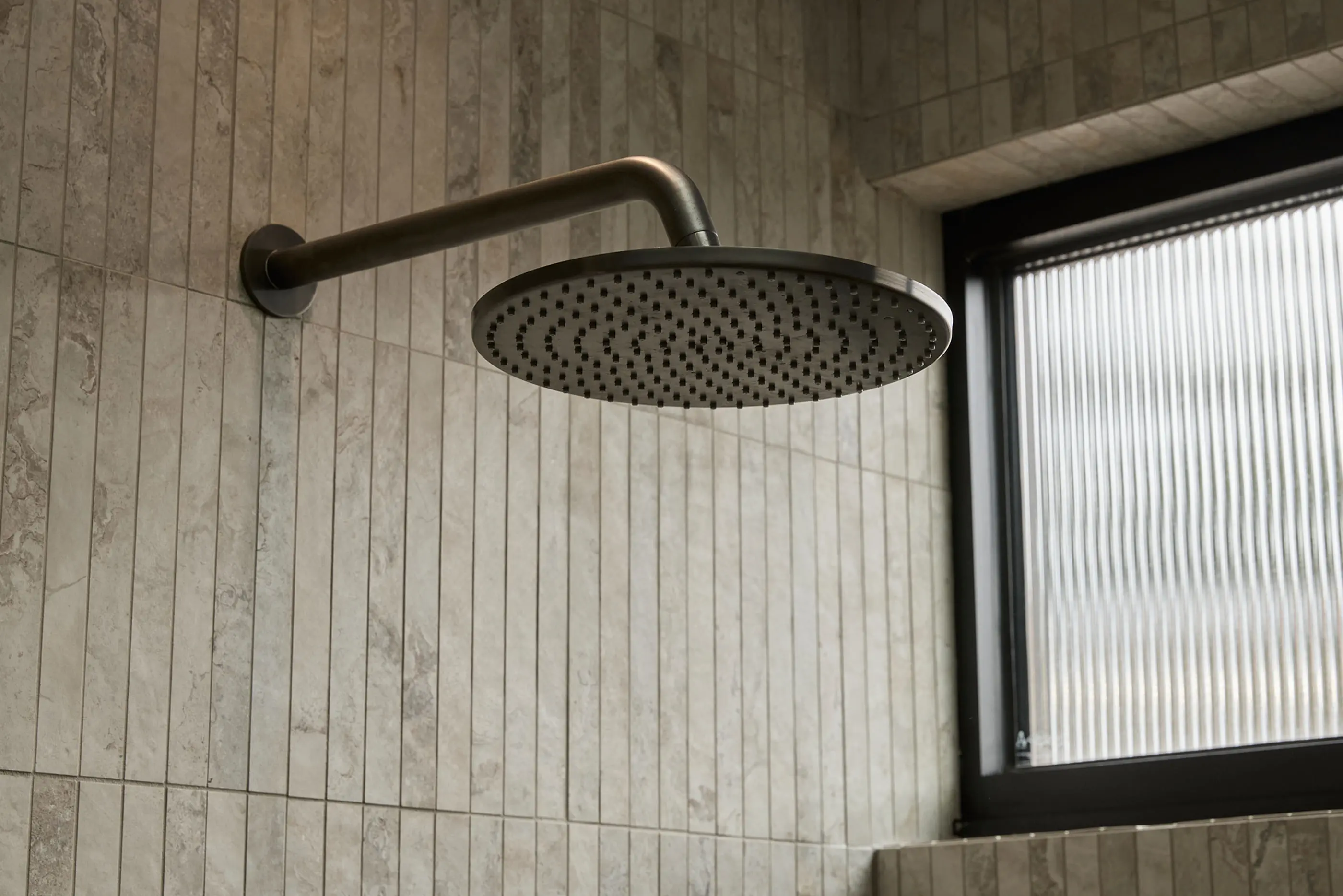 Modern round rainfall showerhead mounted on beige tiled bathroom wall next to a frosted window.