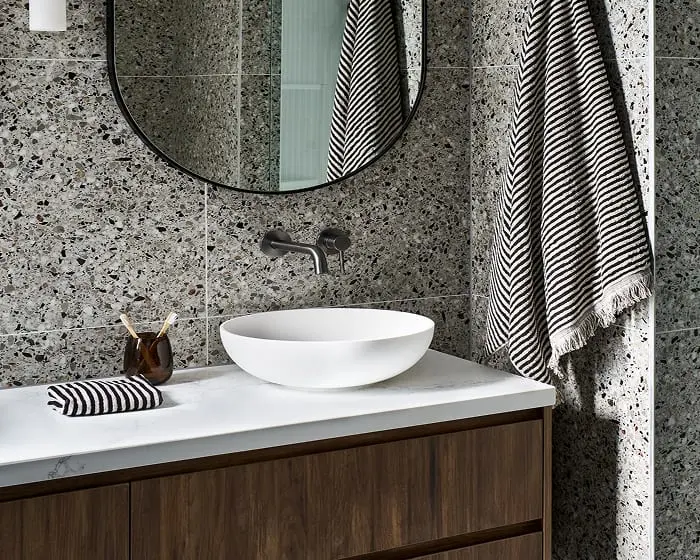 Modern bathroom with terrazzo tile walls, round mirror, white vessel sink, and black-white striped towels.