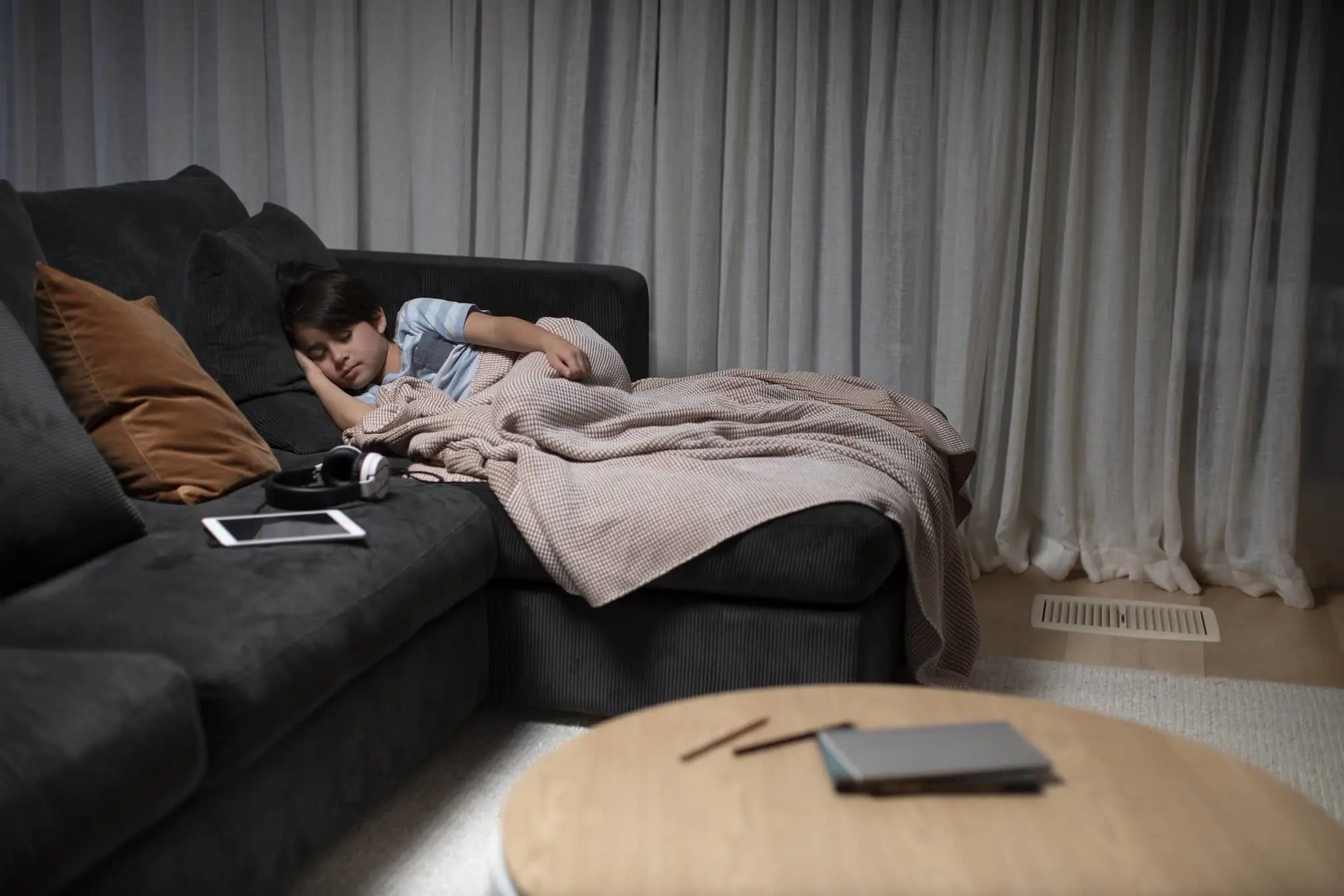 Child sleeping on dark gray couch covered with beige blanket, tablet and headphones nearby in living room with curtains.