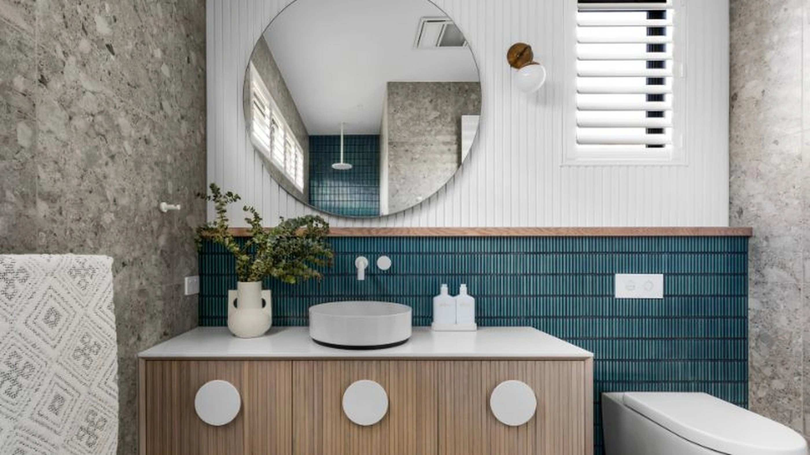 Modern bathroom with round mirror, wooden vanity, teal tile backsplash, concrete walls, and white vessel sink.