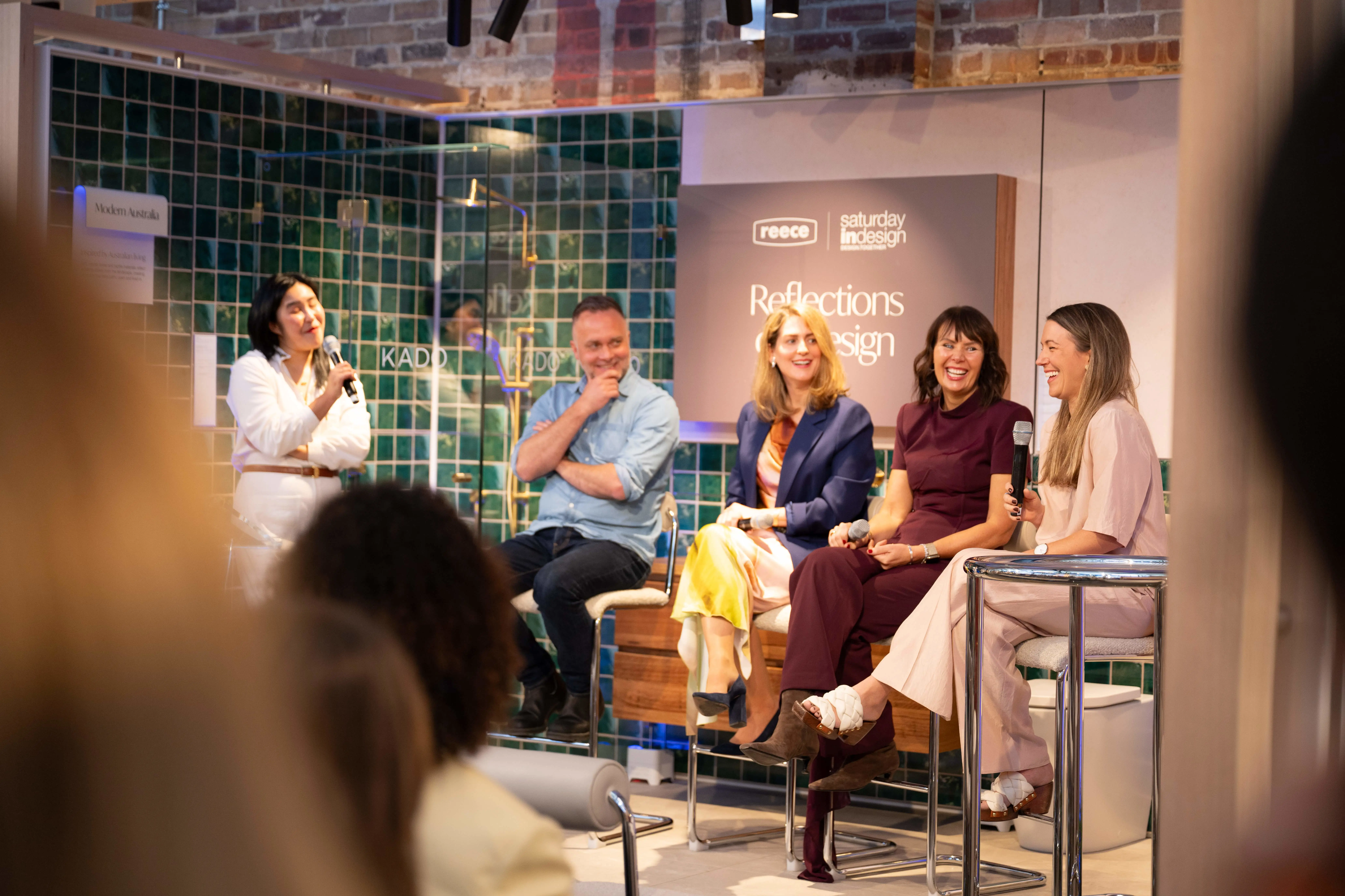 Five people on stage at a panel discussion with green tiled backdrop and Reflections in Design sign visible.