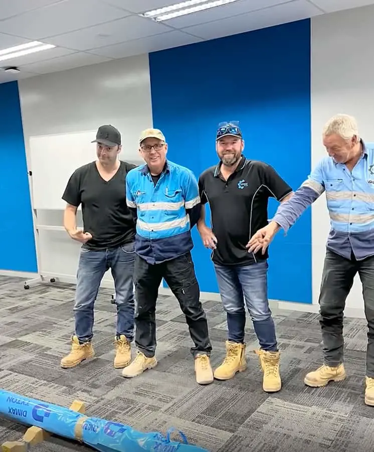 Four workers in work clothes and tan boots standing in an office with blue wall panels and carpeted floor.