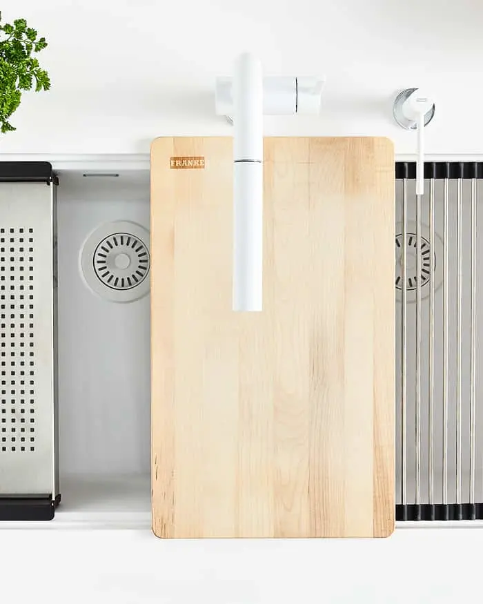 Modern kitchen sink with wooden cutting board, white faucet, metal drain rack, and small plant visible from above.