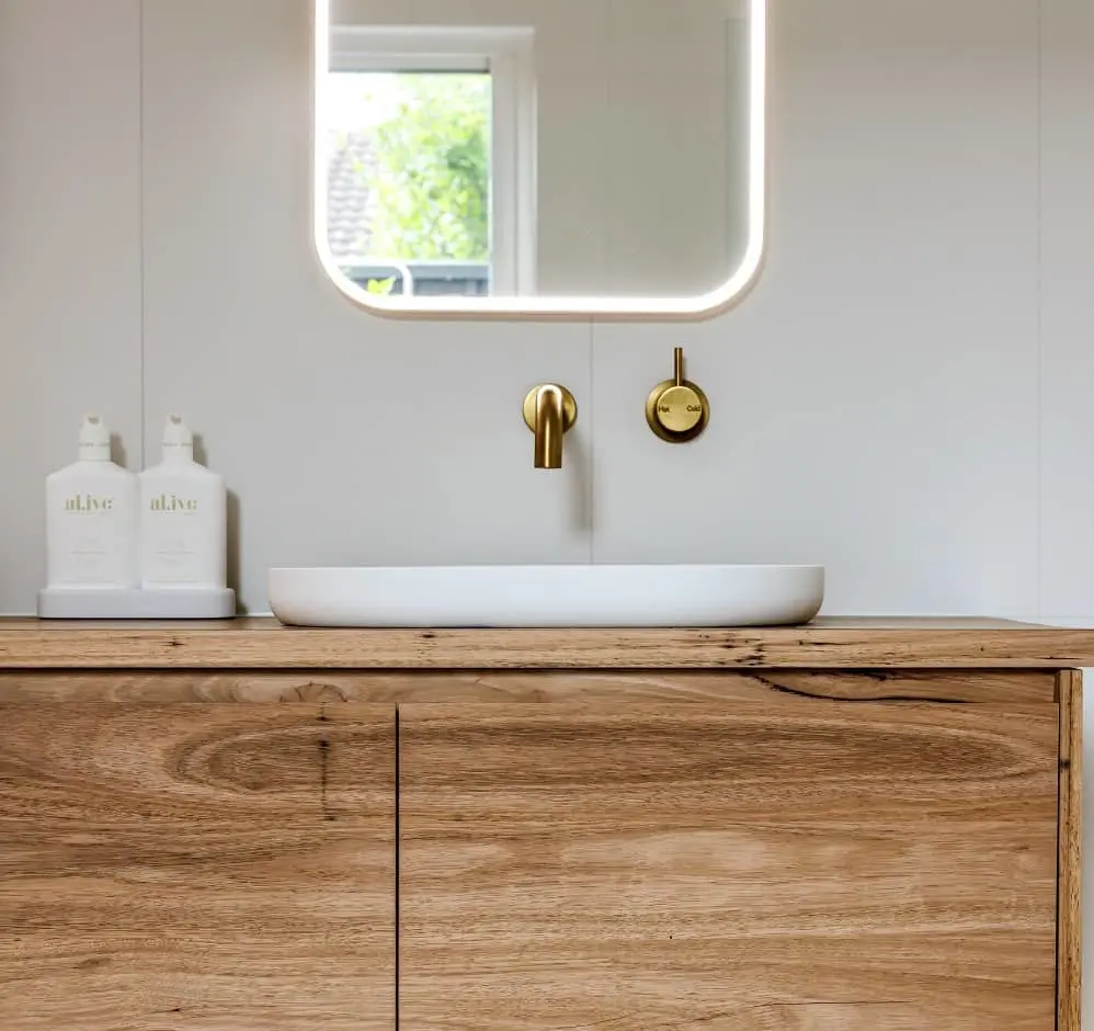 Modern bathroom with white vessel sink on wooden vanity, brass fixtures, illuminated mirror, and white product bottles.