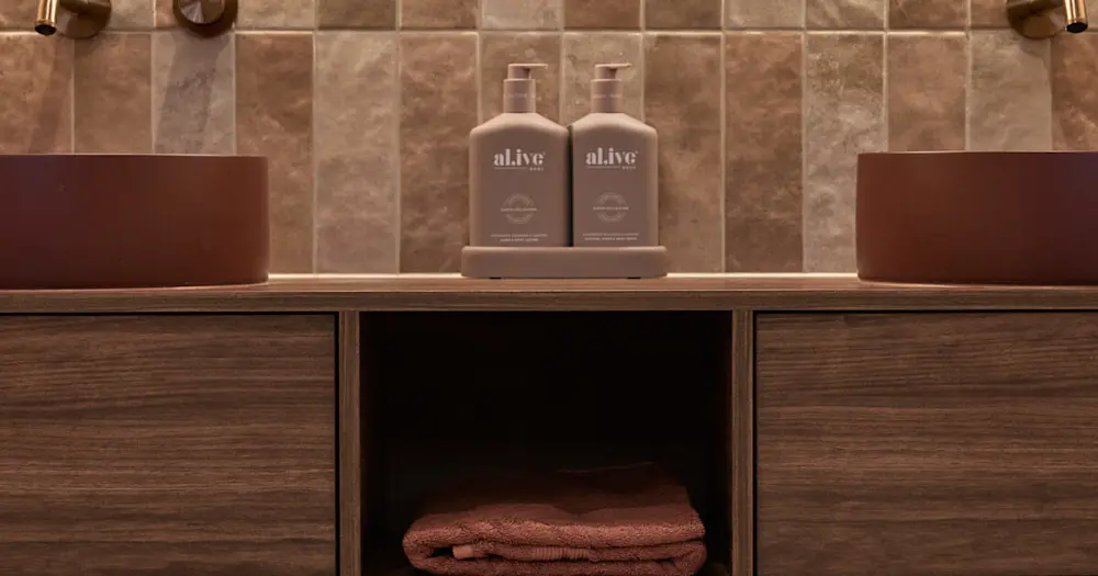 Two "al.ive" brand pump bottles on wooden bathroom vanity with vessel sinks and folded towel in open shelf below.