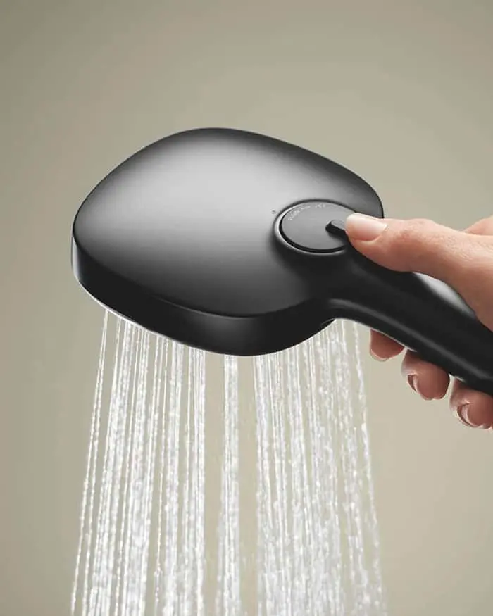 A hand holding a matte black handheld showerhead with a button on top, spraying water downward against a neutral background.
