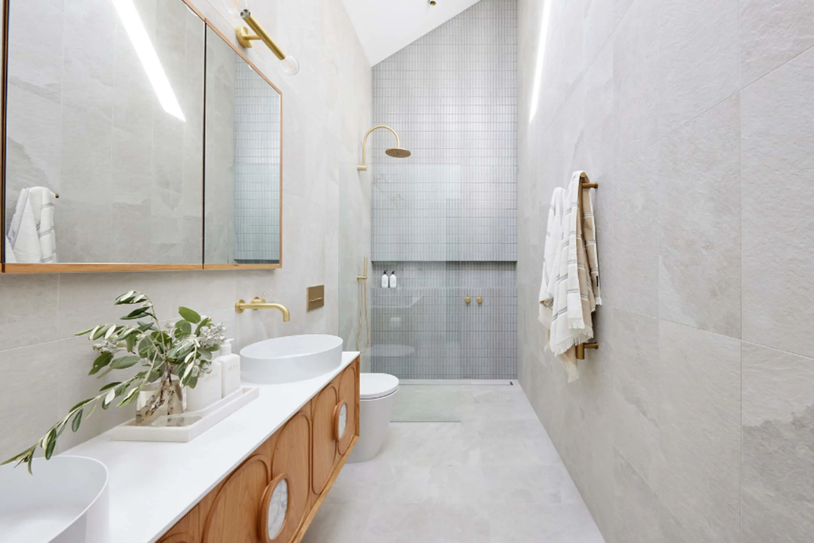 Modern bathroom with white marble tiles, wooden vanity with double sinks, walk-in shower, and gold fixtures.