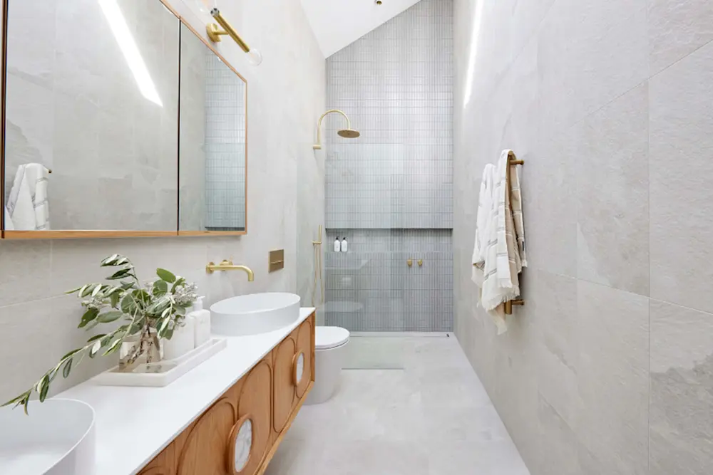 Modern bathroom with white marble tiles, wooden vanity with double sinks, walk-in shower, and gold fixtures.