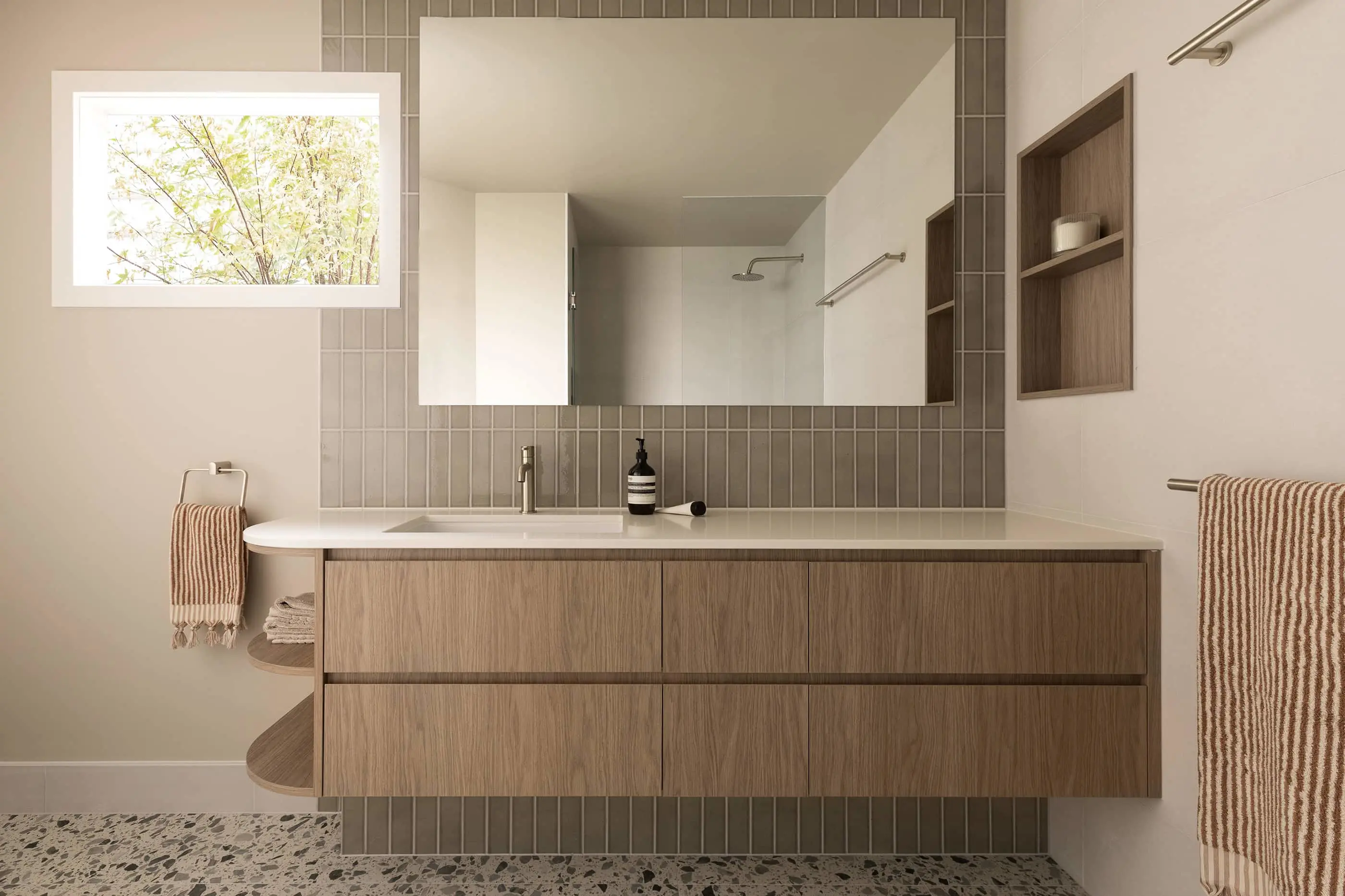 Modern bathroom with wooden vanity, gray tile backsplash, large mirror, and window showing greenery outside.