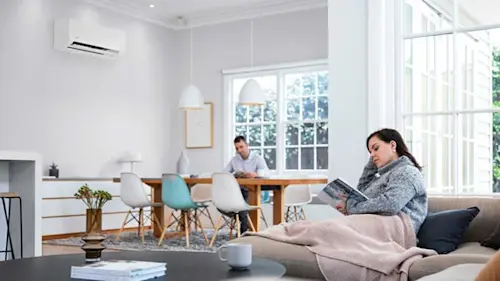 Modern bright living space with woman reading on couch while man works at dining table, air conditioner visible on wall.