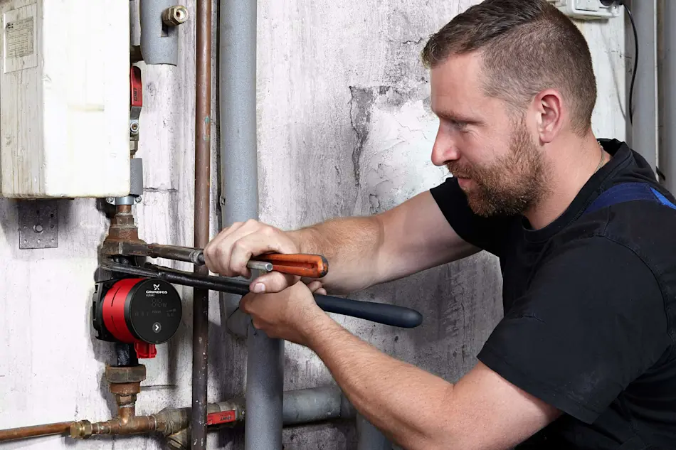 Plumber in dark shirt using wrench to repair water pipes and red Grundfos pump mounted on concrete wall.