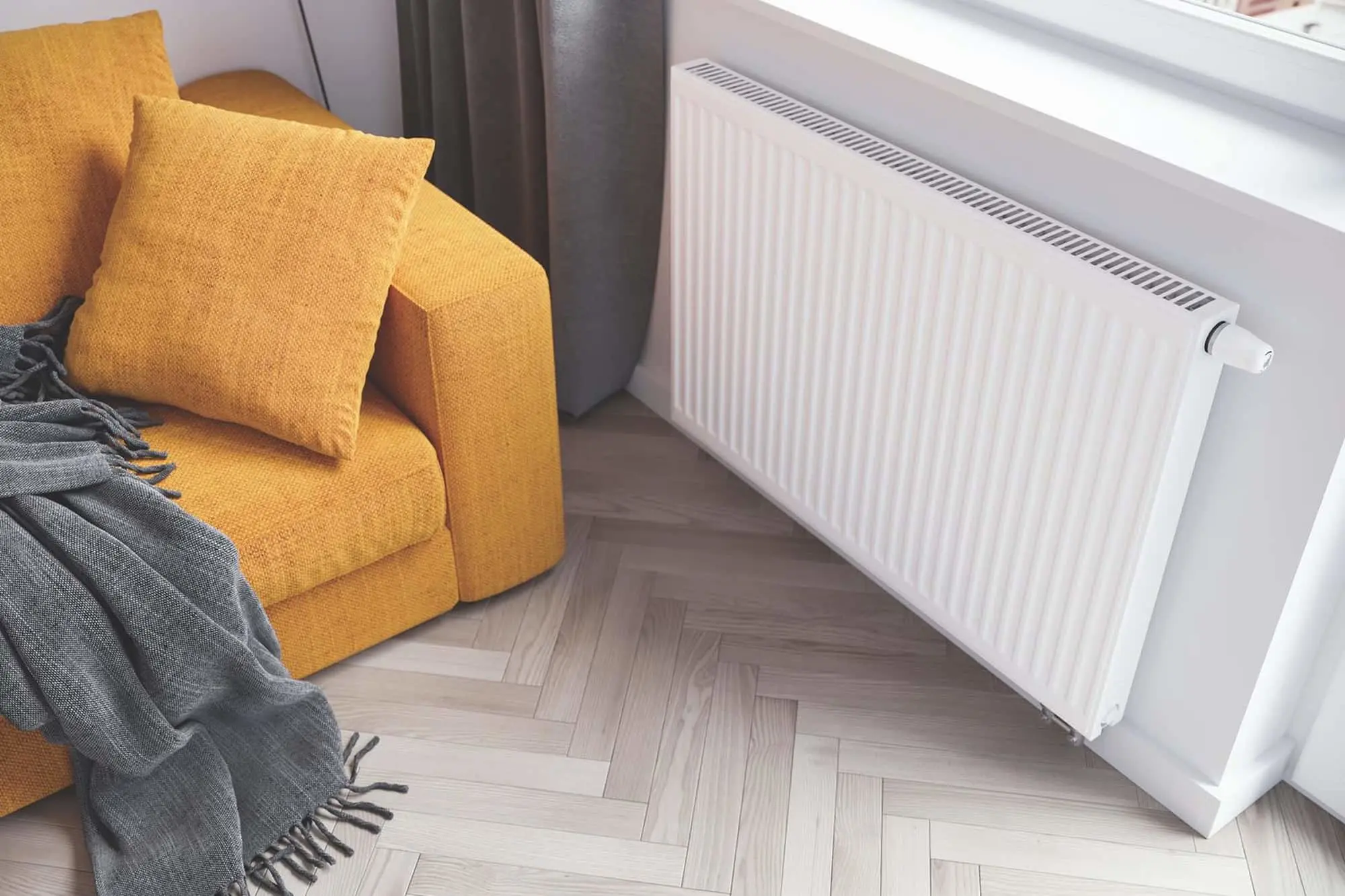 Yellow sofa with cushions and gray throw blanket next to white radiator on herringbone wood floor.