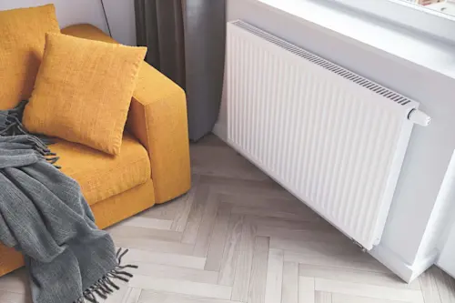 Yellow sofa with cushions and gray throw blanket next to white radiator on herringbone wood floor.