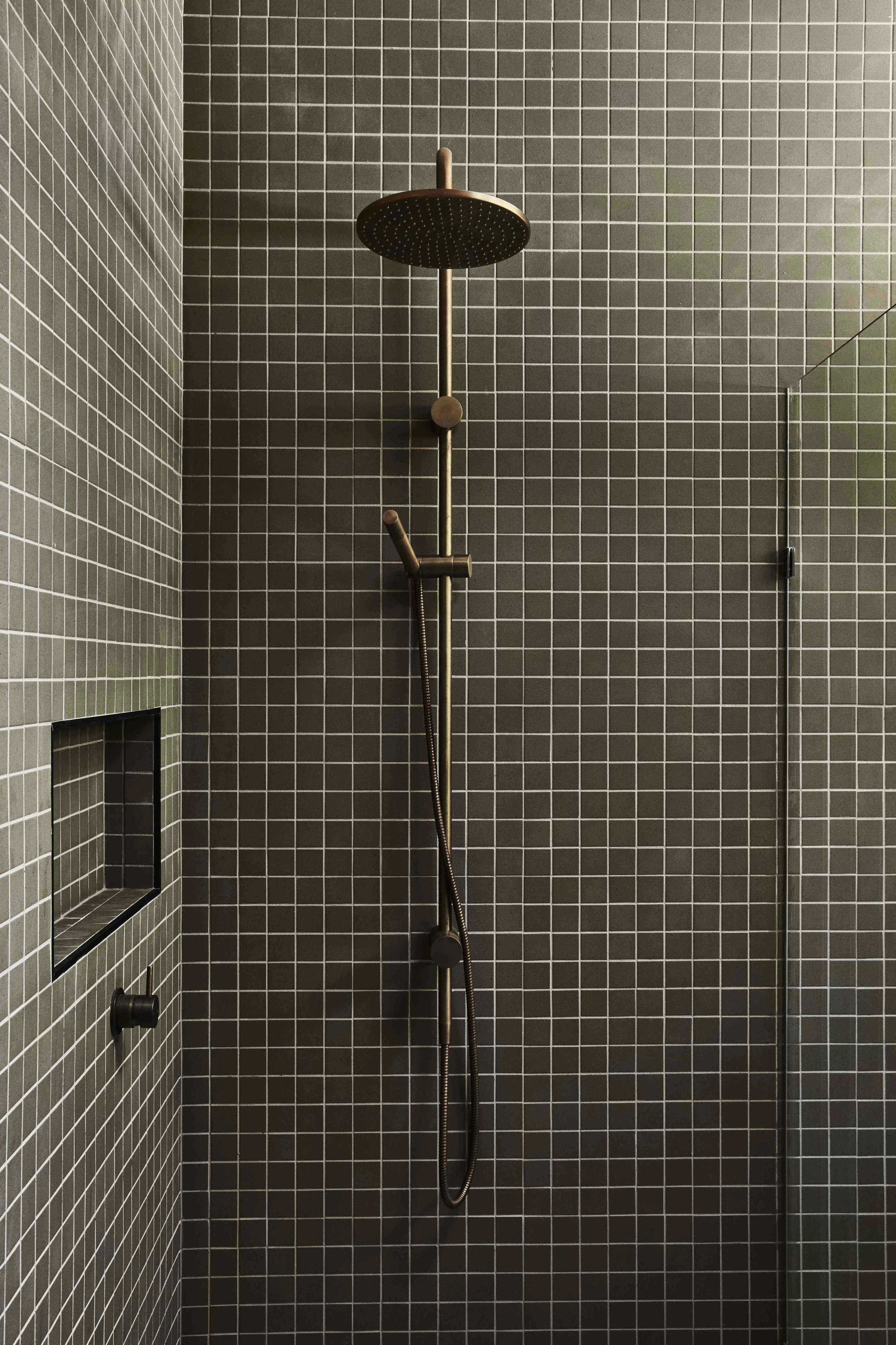 Shower with dark green square tiles, brass rain showerhead, handheld shower on a rail, recessed niche, and glass panel.