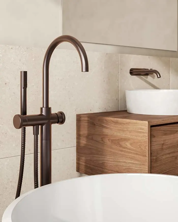 Bronze freestanding bath filler with hand shower next to a white freestanding tub and a wood vanity with vessel sink.
