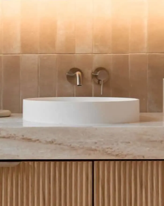 Modern bathroom with white circular vessel sink on marble countertop, wall-mounted faucet, and beige tiled wall.