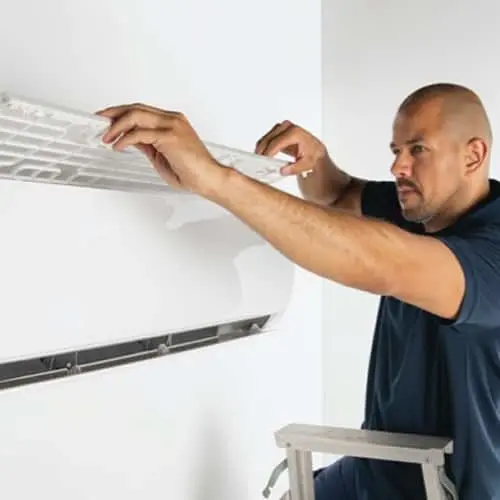 Person in blue shirt installing or servicing a wall-mounted air conditioning unit while standing on a ladder.