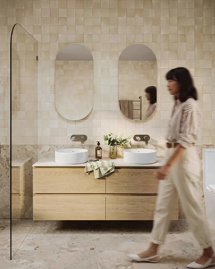 Woman walking past double vanity with oval mirrors and white vessel sinks in tiled bathroom