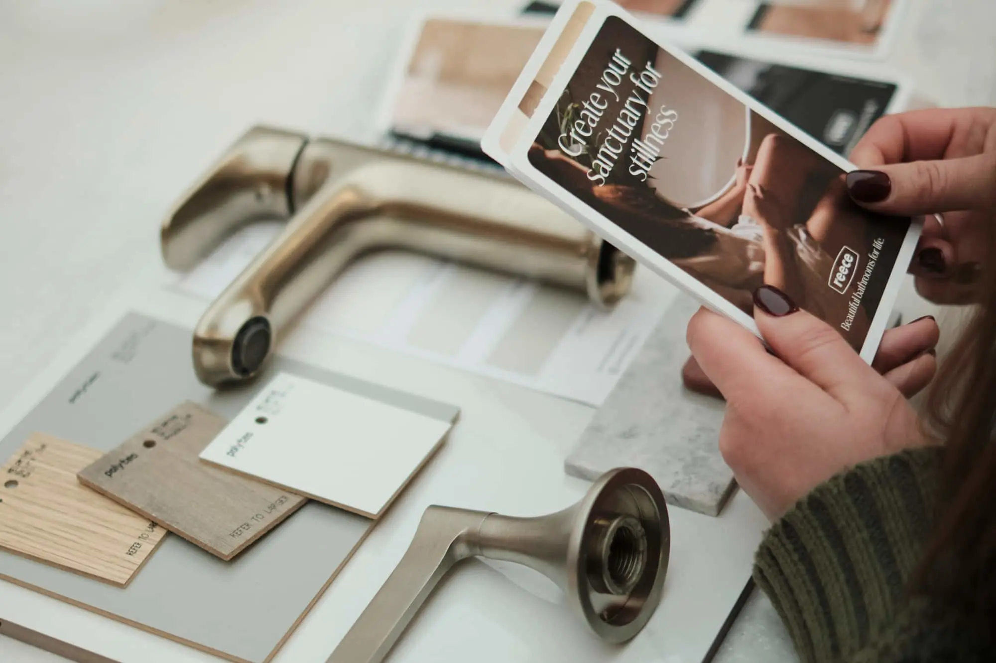 A person holds a Reece brochure over a table with brushed metal tap fittings, wood swatches, and paint color samples.