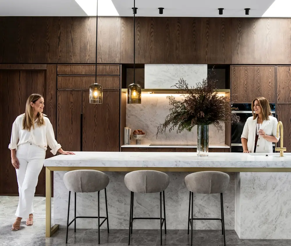 Two women in white outfits in a modern kitchen with marble island, wood cabinetry, pendant lights, and gray barstools.