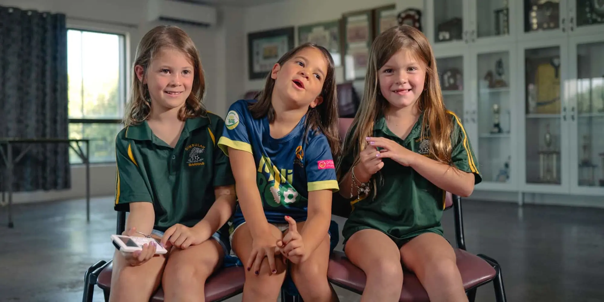 Three children sitting together in a classroom, two wearing green school uniforms and one in a blue sports jersey.