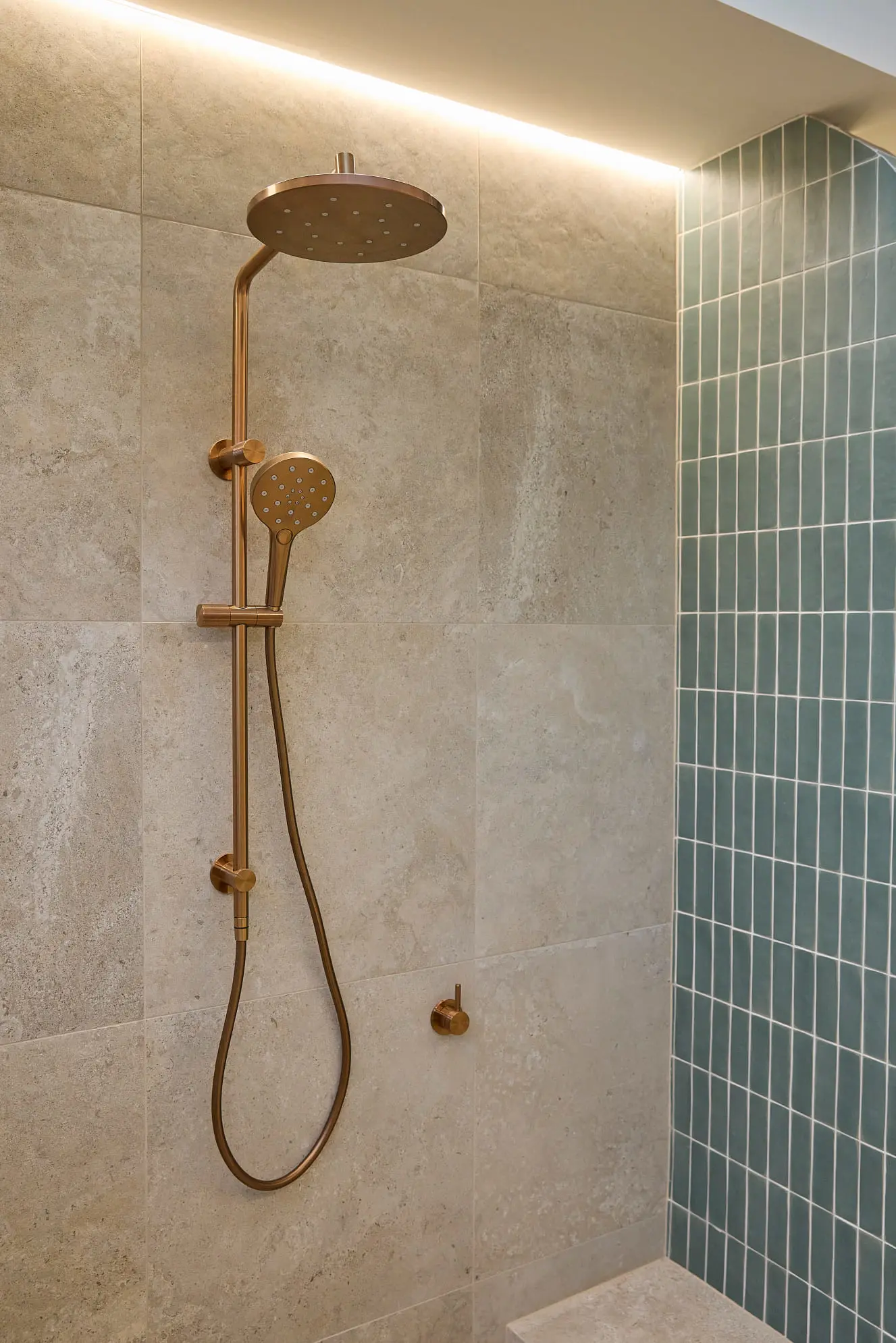 Modern shower with brass rainfall head and handheld sprayer, beige stone tiles and teal accent wall.
