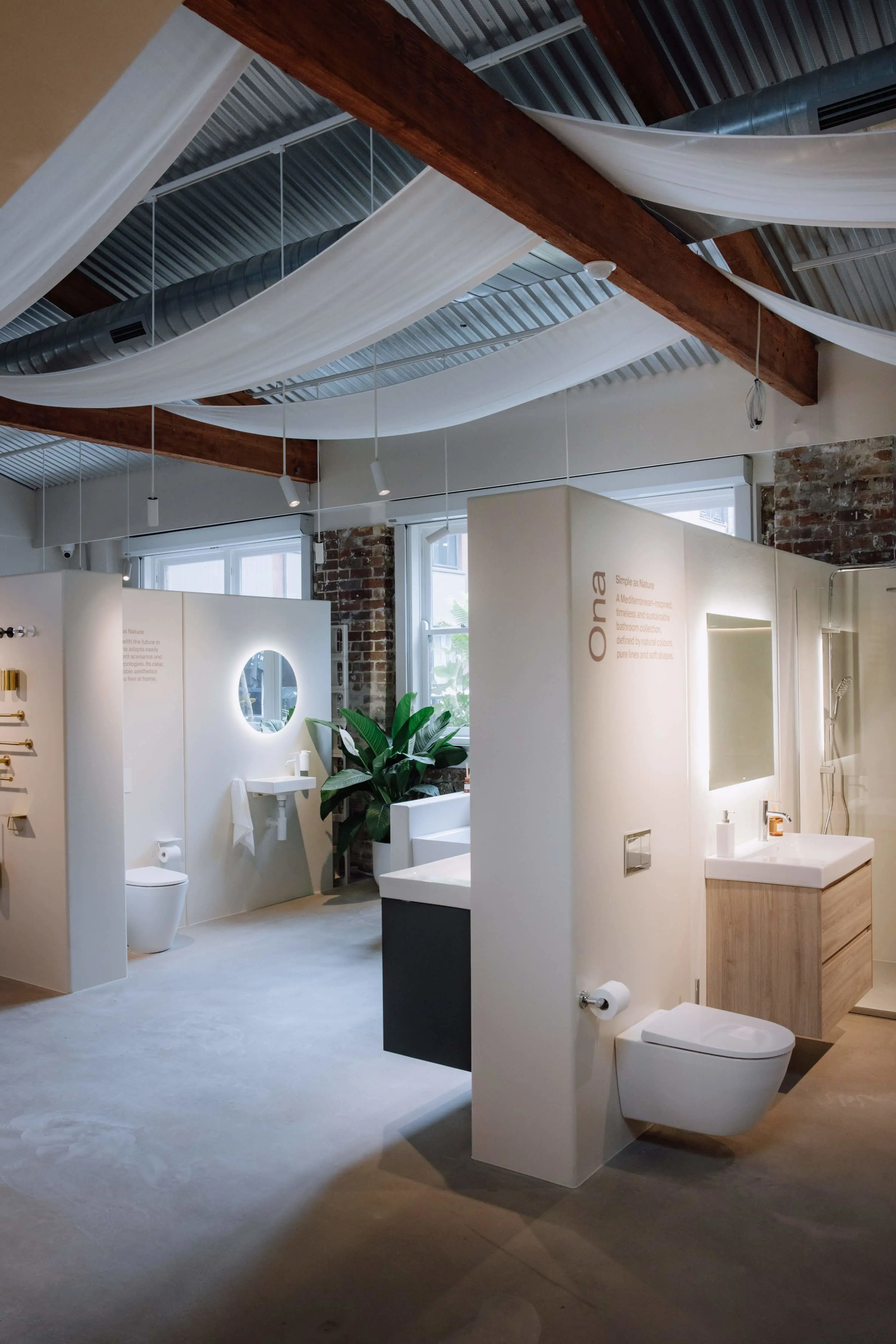 Modern bathroom showroom with white display units, exposed brick walls, wooden beams, and draped fabric ceiling.