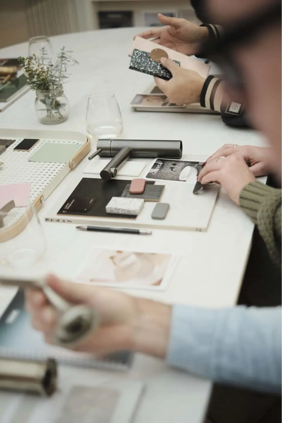 Hands working with material samples and design boards on white table with small plant in jar and empty wine glass.
