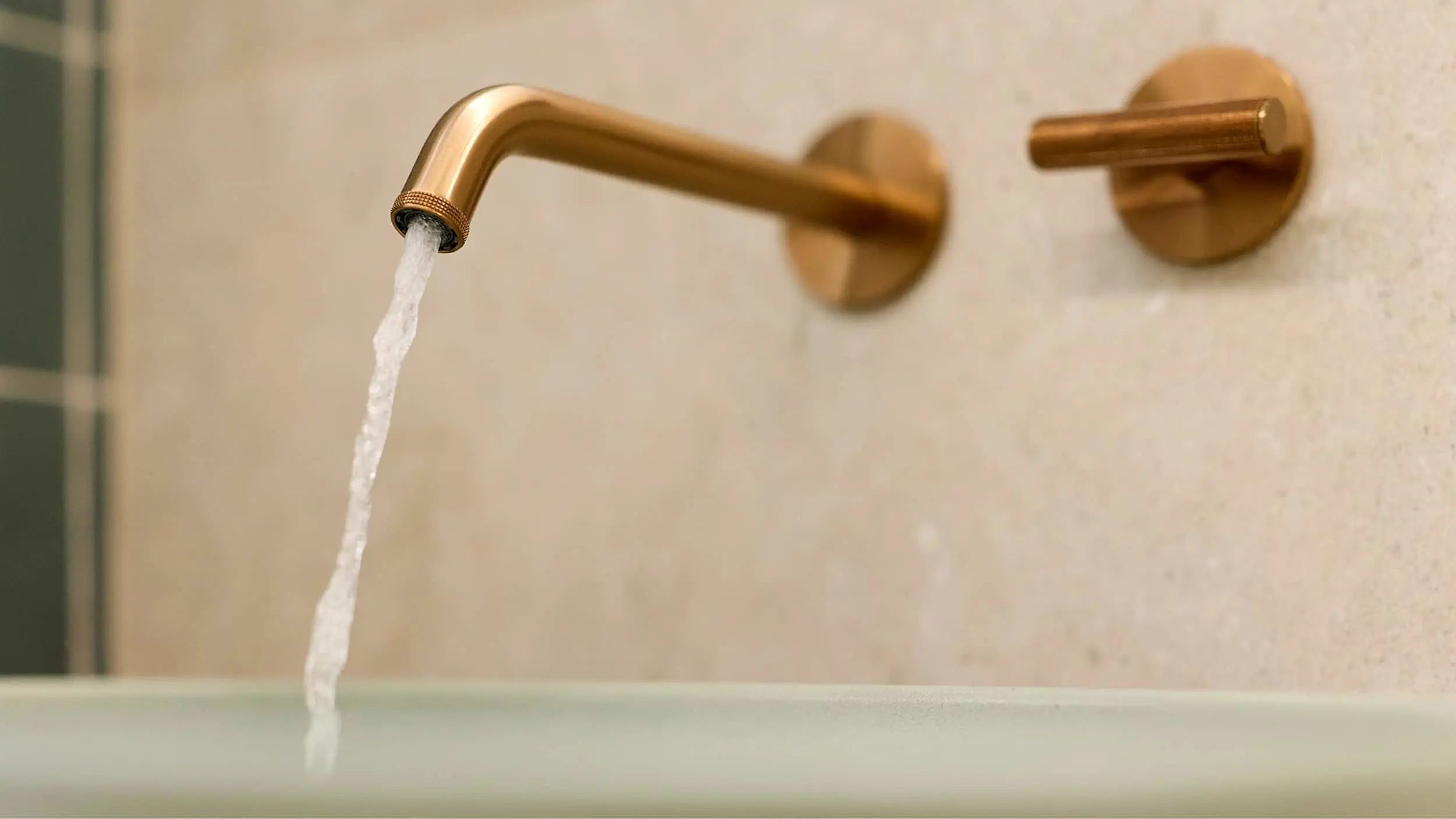 Water flowing from a brass wall-mounted faucet in a bathroom with beige tiled walls.