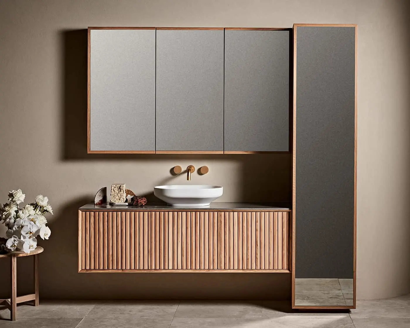 Modern bathroom with wooden vanity, white vessel sink, mirrored cabinet, and tall mirror. White flowers on side table.
