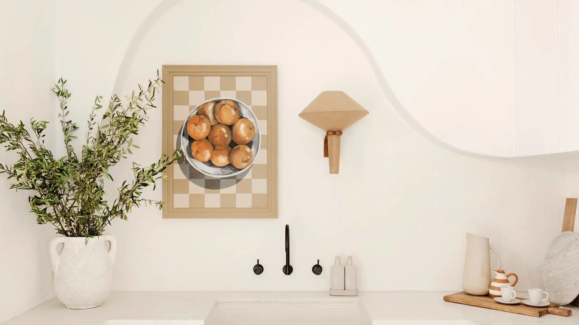 Minimalist kitchen with white walls, potted plant, framed oranges artwork, sink with black fixtures, and ceramic dishware.