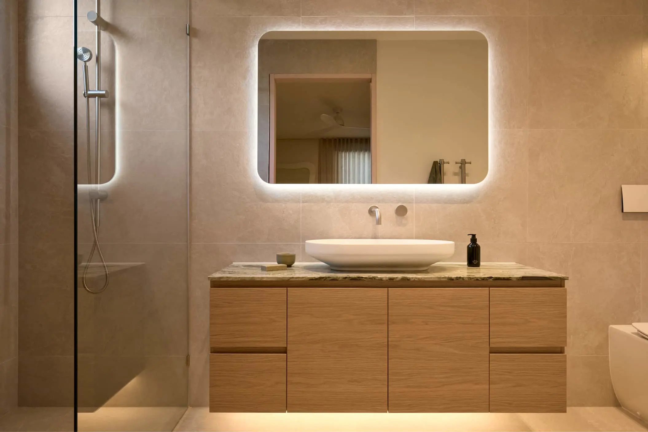 Modern bathroom with beige tiles, backlit mirror, white vessel sink on wood vanity, and glass shower enclosure.