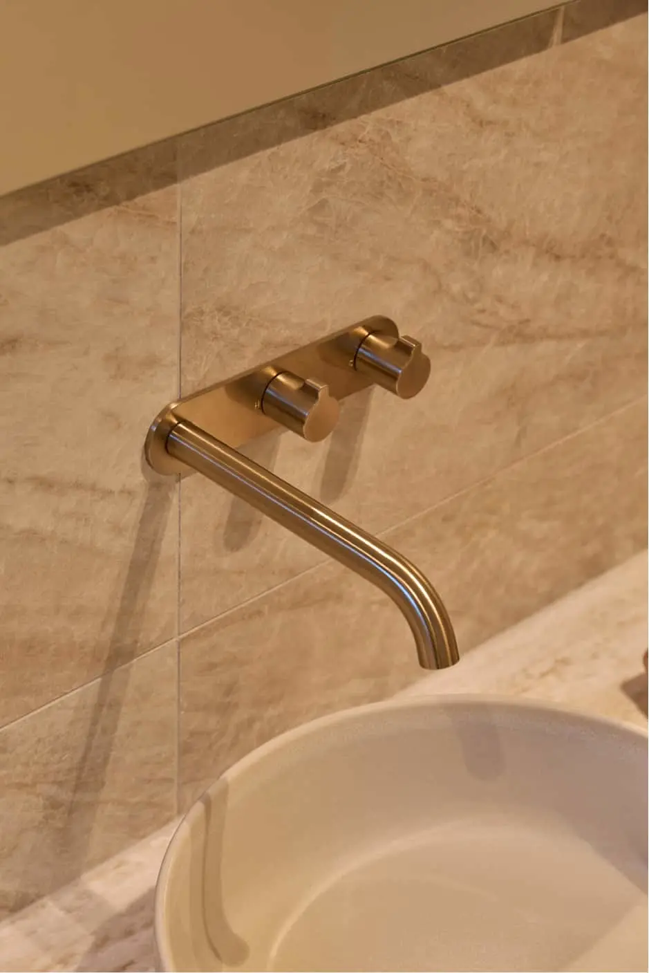 Gold wall-mounted faucet with curved spout above a white sink against beige marble tile wall.