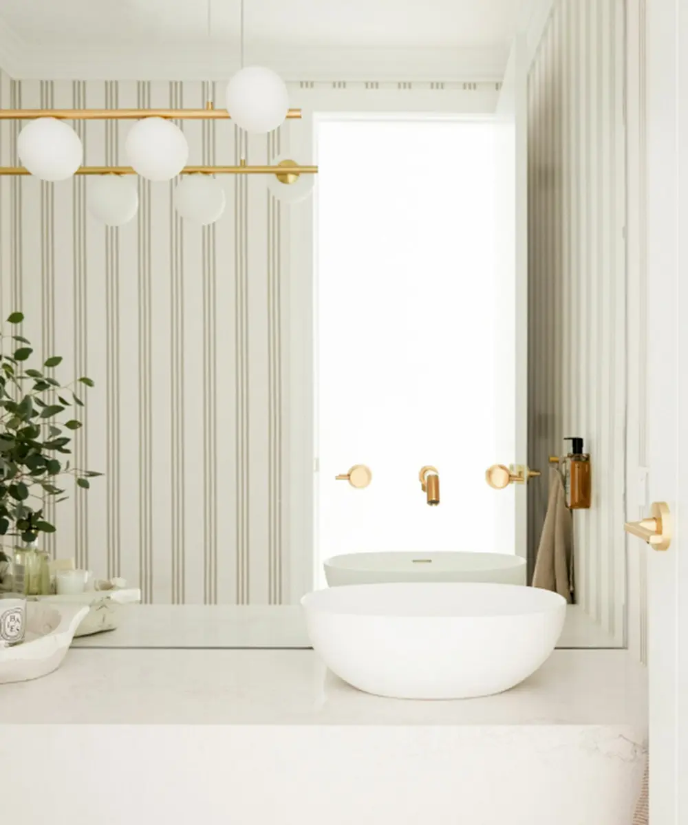 Modern bathroom with striped wallpaper, white vessel sink, gold fixtures, globe lights, and a small plant on the counter.