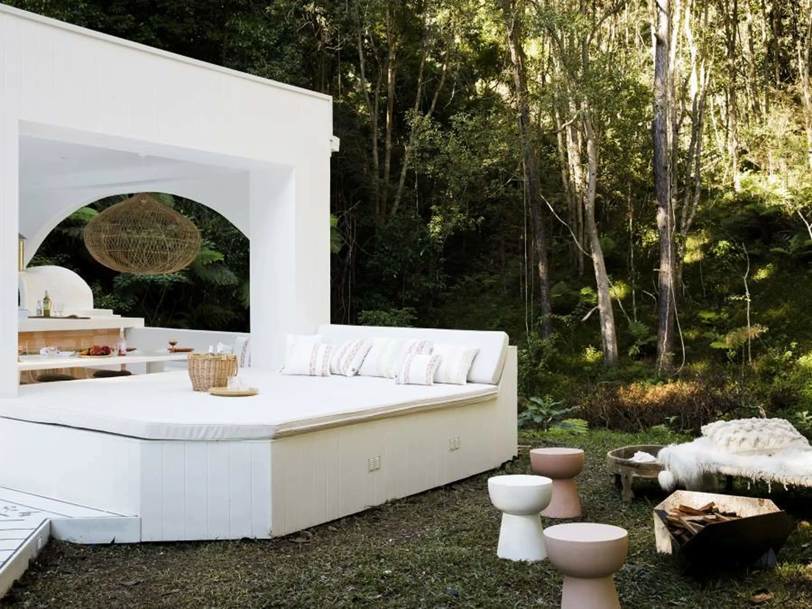 Modern white outdoor lounge area with woven pendant light, surrounded by forest trees and small ceramic stools.