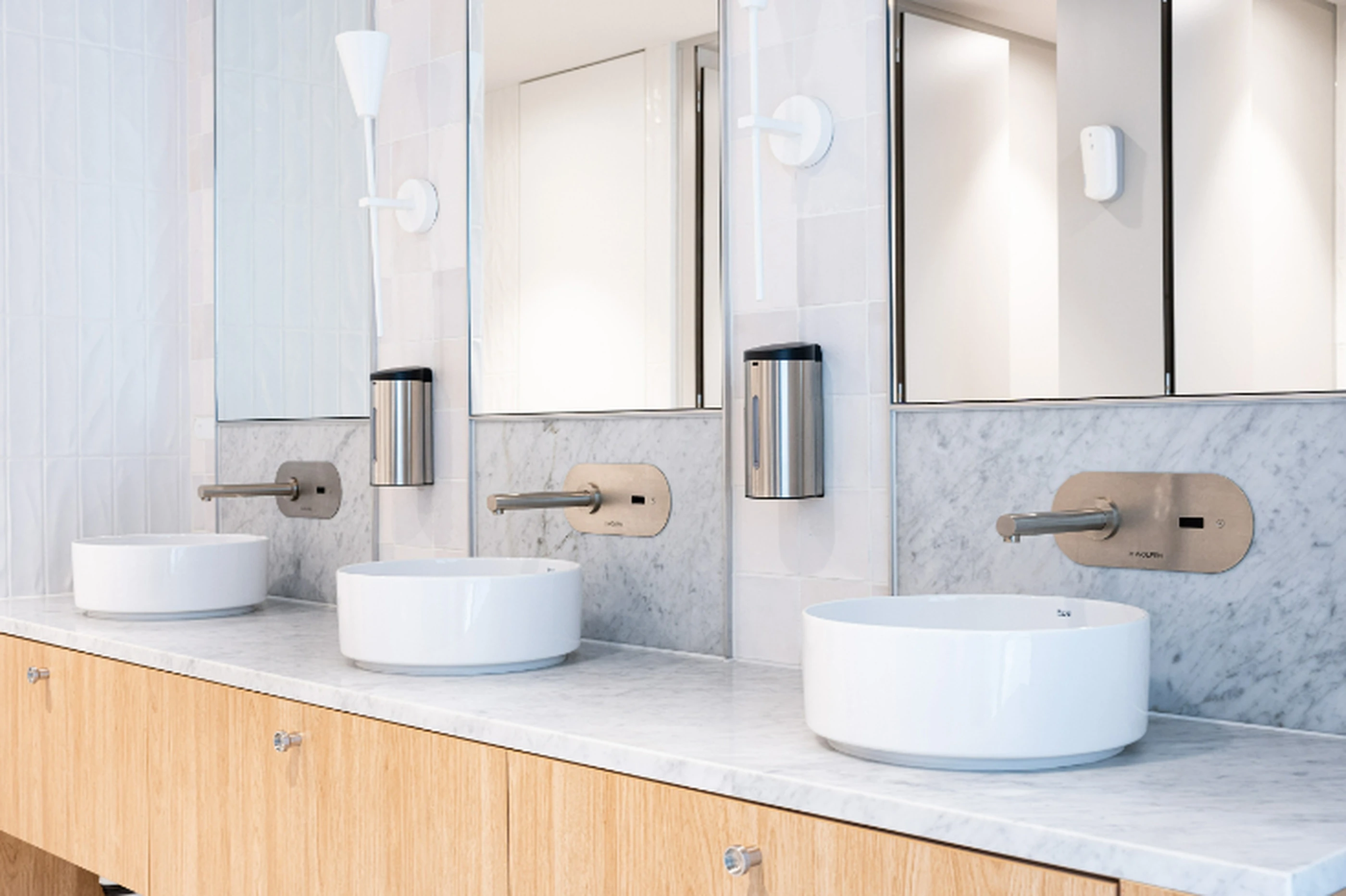 Modern bathroom with three white vessel sinks, wall-mounted faucets, marble backsplash, and mirrored cabinets above.